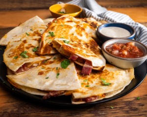 A plate of grilled quesadilla's filled with sliced chicken, crispy Hempler's Bacon and a dipping sauce of ranch dressing in a ramekin.