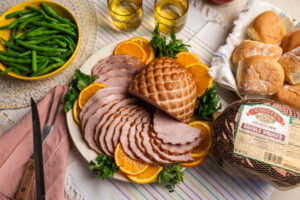 A platter of sliced Hempler’s Double Smoked Ham garnished with fresh parsley and orange slices. The ham is served alongside a bowl of green beans, soft dinner rolls, and a packaged Hempler’s ham in netting. Two glasses of a cold beverage are placed in the background.