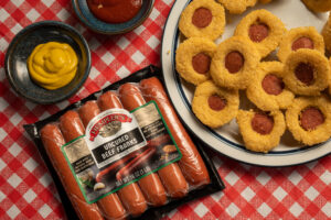 Package of Hempler's hotdogs and a plate of mini corn muffins with Hempler's hot dogs in the middle of each muffin. Served on a red plaid tablecloth.