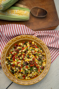 Picnic table setting with a brown bowl full of corn, Hempler's bacon, bell pepper and green onion combined as a salad.