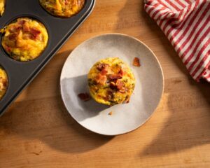 Bacon, egg, and cheddar breakfast muffin topped with crispy bacon on a plate beside a muffin tin filled with baked egg bites.